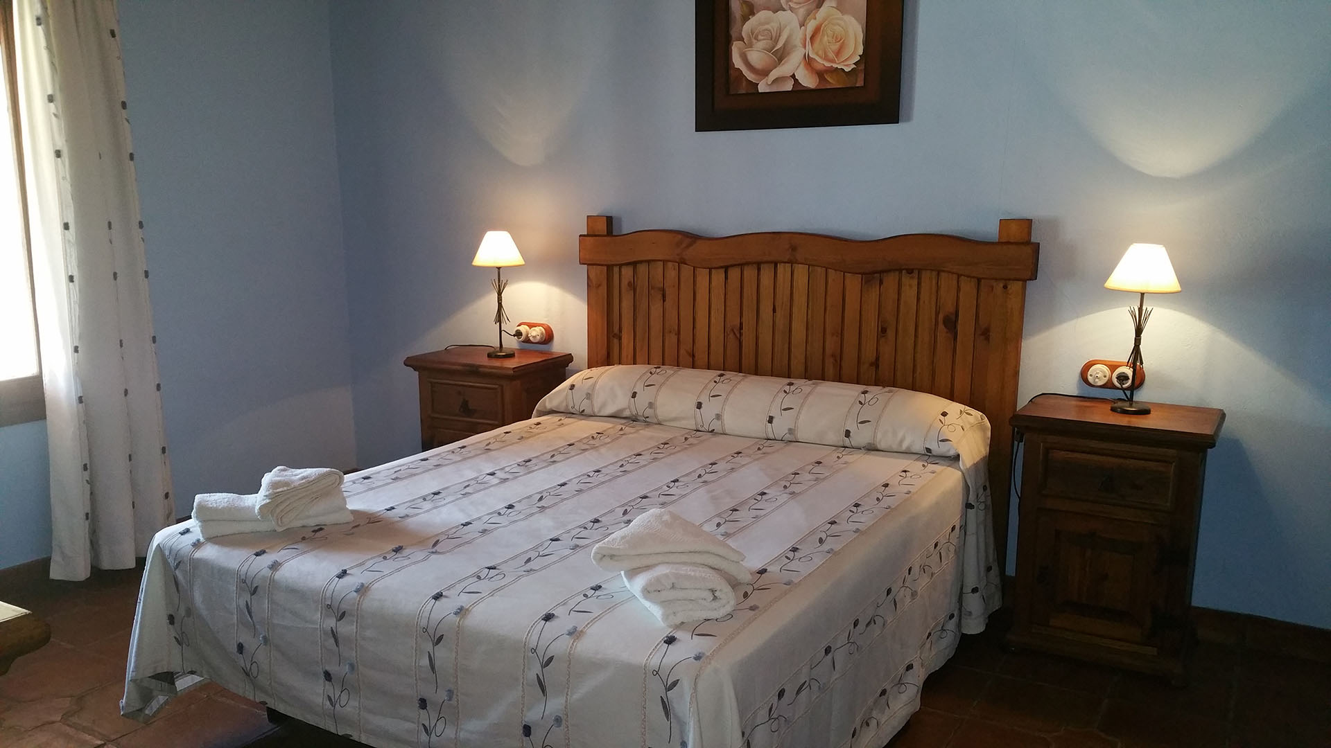 Un dormitorio con cama de matrimonio y dos mesitas de noche con lamparita. Decoración rusticas de madera.