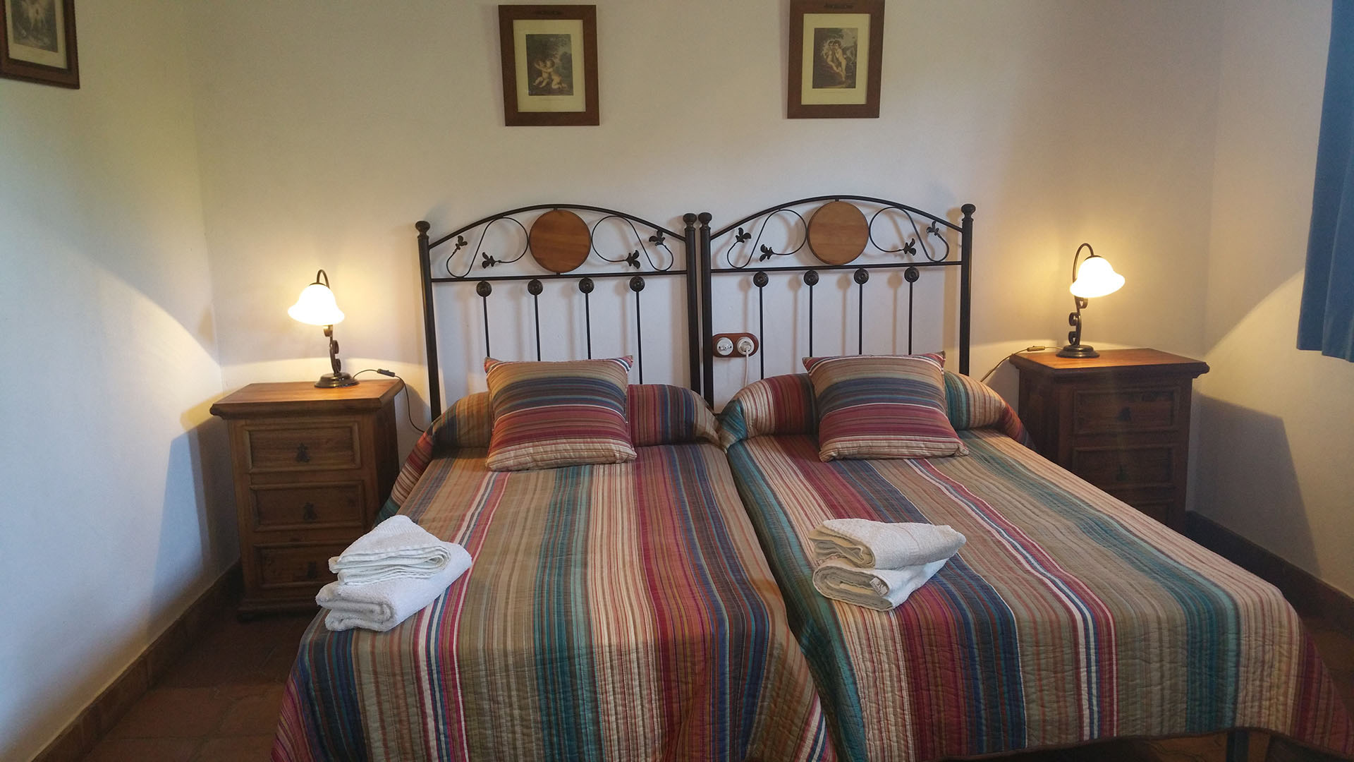 Un dormitorio dos camas individuales y dos mesitas de noche con lamparita. Decoración rusticas de madera y forjado.